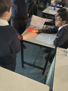 Students at a desk, holding a piece of pink plastic between a piece of paper and a torch